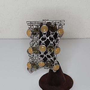 Puede incluir: Una pulsera plateada con un diseño de eslabones de cadena. La pulsera presenta piedras preciosas redondas, amarillo claro y verde oliva alternadas. Se exhibe sobre un soporte de joyería de terciopelo marrón sobre un fondo blanco.