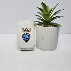 May include: A gold heart-shaped brooch featuring a blue and white butterfly design. The butterfly is set against a blue background with a white moon and palm trees. The brooch has a gold bow at the top.