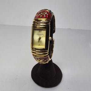 May include: A gold-coloured Joan Rivers Classics quartz watch with a rectangular face and a red and gold enamel band. The watch has a gold dial with black numerals and hands. The watch is displayed on a dark brown stand.