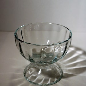 Könnte beinhalten: Eine klare Glasschüssel für Desserts mit geriffeltem Design und einem Sockel. Die Schüssel ist leer und steht auf einer weißen Oberfläche.