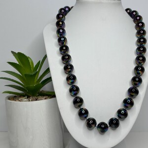 Puede incluir: Un collar de cuentas negras con reflejos iridiscentes se exhibe en un busto de joyería blanco. El collar presenta cuentas grandes y redondas con un acabado brillante. Una pequeña planta en maceta es visible en el lado izquierdo de la imagen.