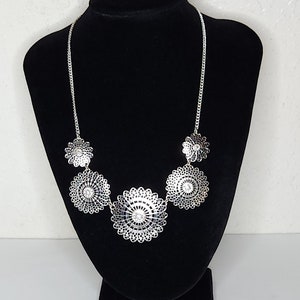 May include: A silver necklace with five large, round, openwork pendants. Each pendant has a smaller circle in the centre with a clear rhinestone.