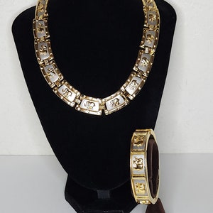 Puede incluir: Un conjunto de collar y pulsera en tono dorado y plateado con un diseño de elefante. El collar está en un soporte de terciopelo negro y la pulsera está en un soporte marrón.