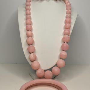 May include: A pink beaded necklace with a large pink bangle bracelet. The necklace is made of large, round beads and has a silver clasp. The bangle is thick and has a smooth surface.