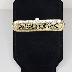 May include: A gold-coloured metal bar brooch with a delicate, intricate design. The brooch has a rectangular shape and is adorned with a pattern of small, open circles and squares.