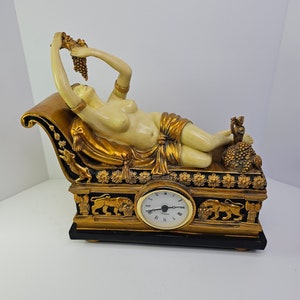 Può includere: Un orologio dorato e nero con una figura scolpita di una donna sdraiata su una chaise longue. La donna tiene un grappolo d'uva e indossa una veste dorata e bianca. Il quadrante dell'orologio è bianco con lancette nere e numeri romani.