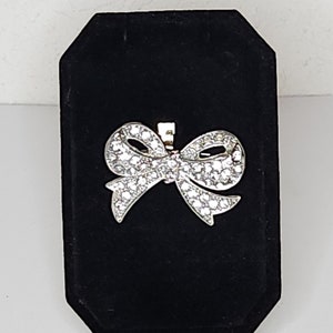 Może przedstawiać: Srebrna broszka w kształcie kokardki z przezroczystymi cyrkoniami. Broszka znajduje się na czarnym aksamitnym stojaku.