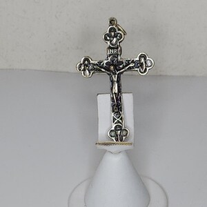 Puede incluir: Un colgante de cruz de plata y oro con detalles intrincados y una figura de Jesucristo en la cruz. El colgante está en un soporte de exhibición blanco.