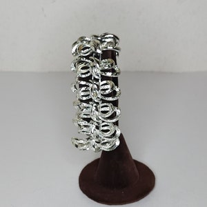 May include: A silver bracelet with a unique design of interlocking loops. The bracelet is displayed on a brown velvet stand.