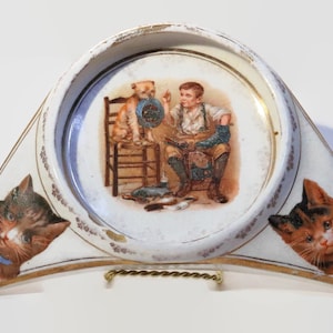Puede incluir: Un cenicero de cerámica blanca con forma de media luna con dos gatos en los extremos y un centro redondo con un niño y un perro. El niño lleva una camisa azul y pantalones marrones y sostiene un zapato. El perro lleva un sombrero azul y está sentado en una silla de madera. El cenicero tiene un borde dorado.