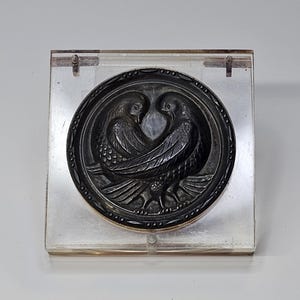 May include: A dark, circular relief carving featuring two birds facing each other, set within a decorative border. The carving is mounted in a clear, square display case. The birds' wings and feathers are detailed, creating a sense of depth.