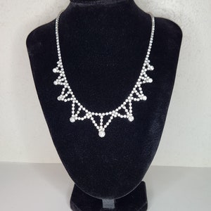 May include: A silver necklace with a geometric design featuring clear rhinestones. The necklace is displayed on a black velvet bust.