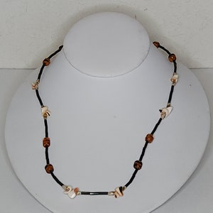 May include: A brown and black beaded necklace with white shell accents. The necklace is displayed on a white mannequin.