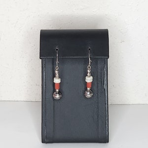 Può includere: Un paio di orecchini pendenti in argento con perline rosse e bianche. Gli orecchini sono esposti su un espositore per gioielli nero.