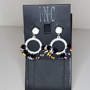 Może przedstawiać: Para srebrnych kolczyków koła z czarnymi i wielokolorowymi koralikami zwisającymi z dołu. Kolczyki są prezentowane na czarnej kartce z nadrukowanym tekstem "I-N-C".
