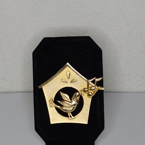 May include: Gold-toned birdhouse brooch with a bird singing a musical note. The birdhouse is on a black velvet background.