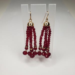 May include: A pair of red crystal tassel earrings with gold accents. The earrings are hanging from a clear acrylic display stand.