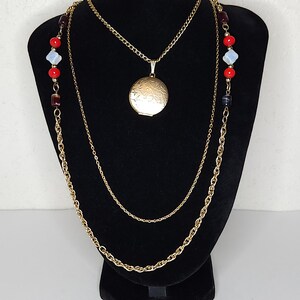 Op de afbeelding: Drie gouden kettingen met een ronde hanger. De kettingen zijn op een zwarte standaard gelegd. De hanger heeft een bloemmotief. De kettingen zijn versierd met rode en witte kralen.