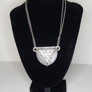 May include: A silver necklace with a geometric pendant. The pendant is a triangle with a textured surface and a rounded bottom.