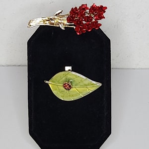 May include: Two gold tone brooches on a black velvet display stand. The first brooch is a cluster of red flowers. The second brooch is a green leaf with a red ladybug.