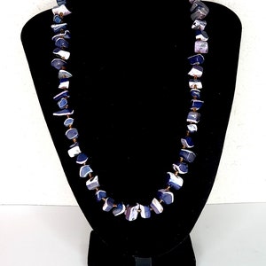 Puede incluir: Un collar largo hecho de cuentas con patrones de color morado, blanco y gris. El collar se muestra en un busto de maniquí de terciopelo negro.