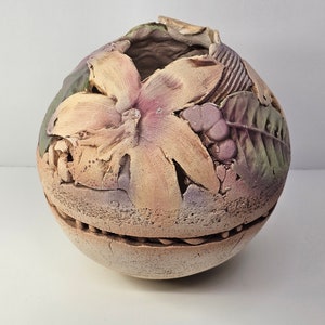 May include: A round ceramic vase with a textured surface and a floral design. The vase is decorated with a large flower, leaves, and berries in shades of pink, yellow, green, and brown.