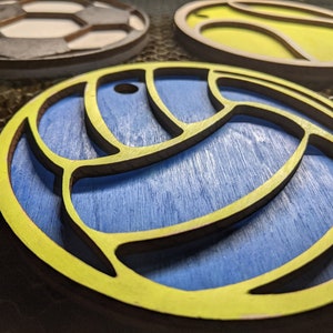May include: A yellow wooden cutout of a volleyball with a blue background. The volleyball has a detailed design with a raised edge.