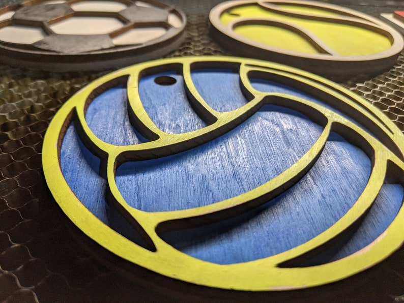May include: A yellow wooden cutout of a volleyball with a blue background. The volleyball has a detailed design with a raised edge.