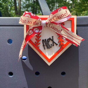 May include: A wooden baseball home plate with a red border and a white background. The plate has a red and white ribbon tied around it with the text "Out to the Ball Game!" printed on the ribbon. The plate also has the name "Alex" written on it in black.