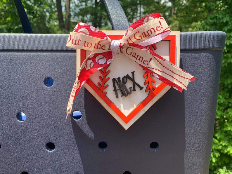May include: A wooden baseball home plate with a red border and a white background. The plate has a red and white ribbon tied around it with the text "Out to the Ball Game!" printed on the ribbon. The plate also has the name "Alex" written on it in black.