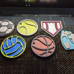 May include: Six wooden ornaments shaped like different sports balls and a pair of ballet shoes. The ornaments are painted in various colors, including white, black, red, blue, yellow, and brown.