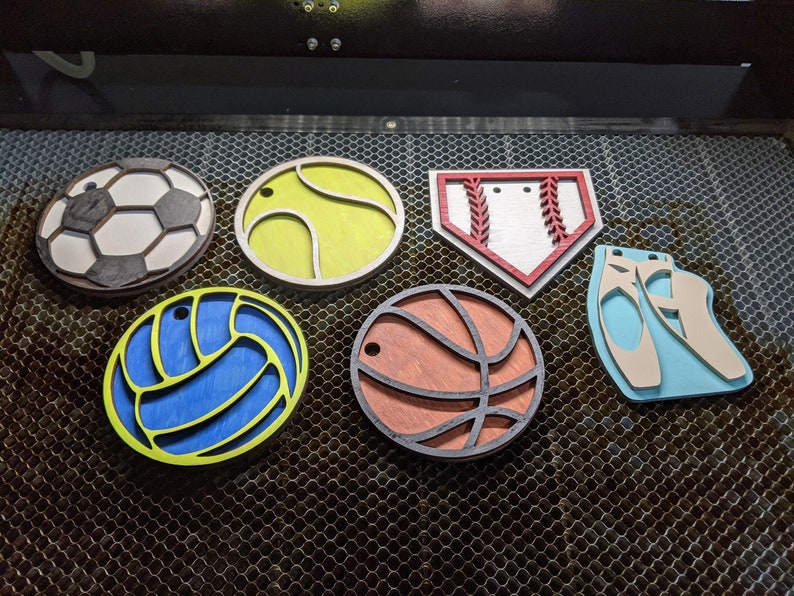 May include: Six wooden ornaments shaped like different sports balls and a pair of ballet shoes. The ornaments are painted in various colors, including white, black, red, blue, yellow, and brown.