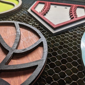 May include: Four wooden cutouts of different sports, including a baseball, basketball, football, and soccer ball. The cutouts are painted in various colors, including red, white, blue, green, and black.