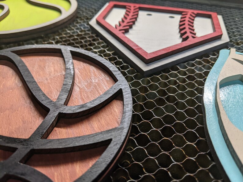 May include: Four wooden cutouts of different sports, including a baseball, basketball, football, and soccer ball. The cutouts are painted in various colors, including red, white, blue, green, and black.