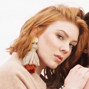 May include: Two women with red and blonde hair, one wearing a beige coat and large tassel earrings with gold accents. The earrings are made of beige and red yarn.