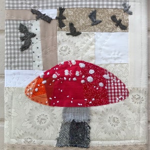 May include: A red and white patchwork mushroom with a grey fabric stem, surrounded by a white background with a grey and white gingham pattern and small grey birds flying above.