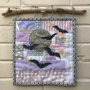 May include: A handmade patchwork quilt featuring a grey moon with bats flying around it. The quilt is made of various shades of purple, pink, and grey fabrics with a black and white border. The quilt is hung on a wooden branch.