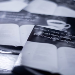 Op de afbeelding: Een stapel zwart-witte pagina's met een bijbelvers gedrukt op de bovenste pagina. De tekst luidt: "JEZUS ANTWOORDDE: "ER STAAT GESCHREVEN: 'DE MENS ZAL NIET ALLEEN VAN BROOD LEVEN, MAAR VAN ELK WOORD DAT UIT DE MOND VAN GOD KOMT.' Mattheüs 4:4"