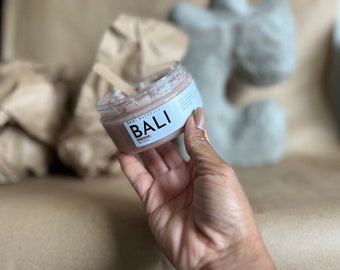 May include: A clear plastic jar containing a light pink body scrub with a wooden applicator stick inside. The white label features the text Demi Naturals BALI body scrub 8oz 227ml. The jar is held up, showcasing the granular texture of the pink product.