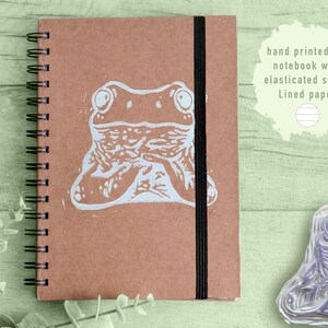 Peut inclure: Un carnet A6 marron avec une impression de grenouille blanche et une sangle élastique noire. Le carnet a une reliure à spirale et comprend le texte "hand printed A6 notebook with elasticated strap. Lined paper."