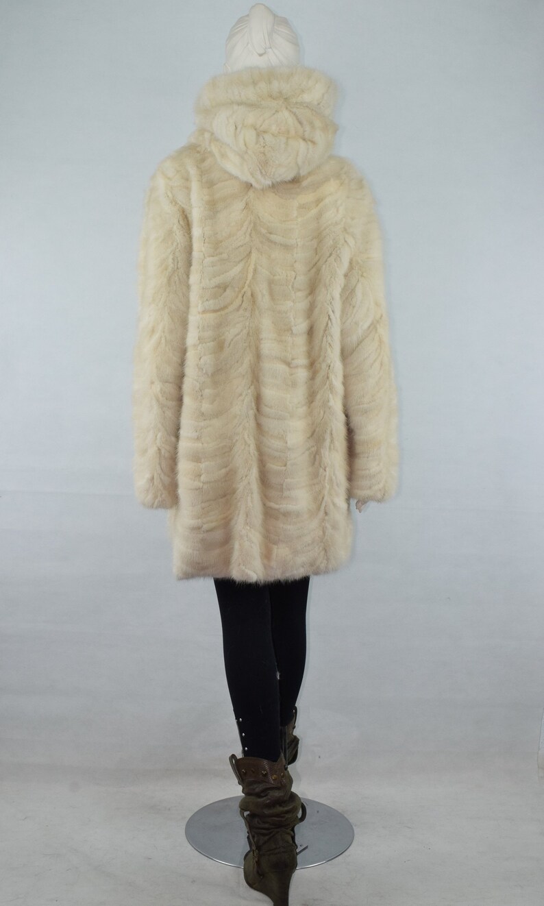 May include: A white fur coat with a hood. The coat is made of a soft, fluffy fur and has a long, flowing style. The coat is being worn by a mannequin.