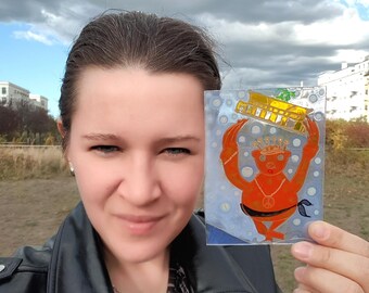 May include: A woman holds a clear plastic sheet with a hand-painted illustration of a red bear wearing a crown and a peace sign necklace. The bear is holding a yellow building above its head.