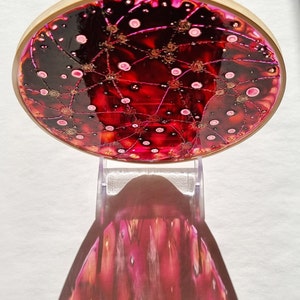 May include: A circular piece of art with a red, black, and gold design. The art is framed in a wooden embroidery hoop and is on a clear stand.