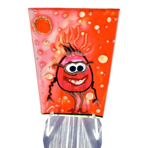 Könnte beinhalten: Eine rote Glasfliese mit einer Comicfigur, die eine Brille trägt und ein breites Lächeln zeigt. Der Hintergrund ist ein rot-weißes, gesprenkeltes Design.