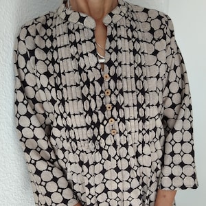 Op de afbeelding: Een zwart-wit gepatroneerde tuniek met een knoopsluiting aan de voorkant en lange mouwen. Het patroon heeft kleine cirkels met een gestreept ontwerp.