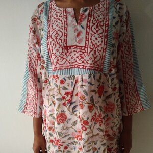 May include: A white cotton tunic with a floral print in shades of red, pink, and green. The tunic has a V-neckline and a gathered waist. The sleeves are three-quarter length and have a blue and red border.