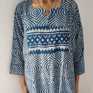 Pode incluir: Uma túnica com padrão azul e branco com decote em V e mangas 3/4. A túnica tem um design geométrico com linhas brancas sobre um fundo azul.