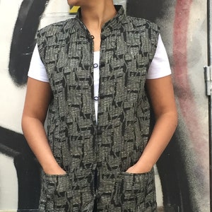May include: A long black and silver patterned vest with a mandarin collar and two pockets. The vest has a button closure.