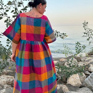 May include: A colorful, plaid-patterned dress with short sleeves. The dress features a vibrant mix of colors including pink, orange, blue, and purple. The dress is worn outdoors near a body of water.