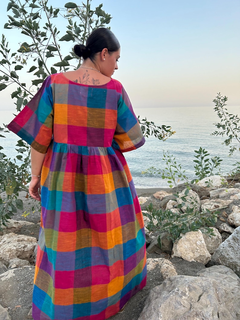 May include: A colorful, plaid-patterned dress with short sleeves. The dress features a vibrant mix of colors including pink, orange, blue, and purple. The dress is worn outdoors near a body of water.